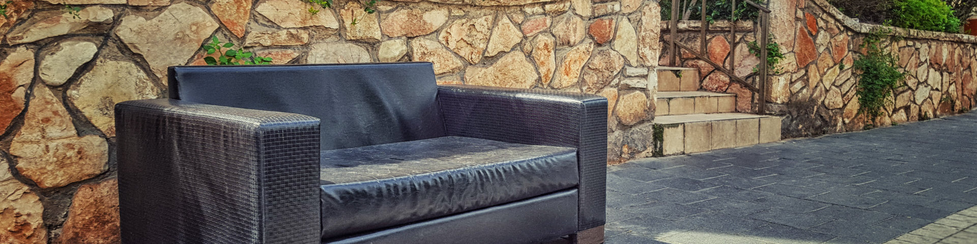 Black leather wooden sofa is dumped on pavement street. This used couch is placed on decorative stone wall background. Bright sunny day